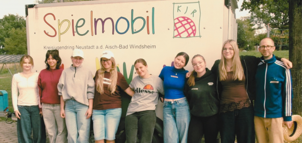 Gruppenbild mit neun ehrenamtlichen Mitarbeitenden des Spielmobils vor dem Spielmobil-Anhänger